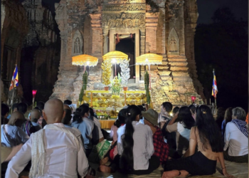 De nuit, au temple de Lolei dans la province de Siem Reap, un groupe de fidèles assis sur des nattes fait face à un ancien sanctuaire en briques illuminé, décoré de parasols jaunes, de fleurs et d’offrandes, en attendant le moment précis fixé par la position de la lune pour lancer une cérémonie de la Fête de l’Eau.