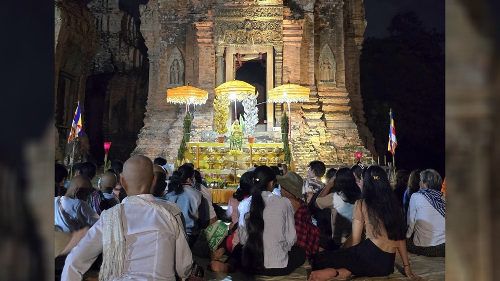 De nuit, au temple de Lolei dans la province de Siem Reap, un groupe de fidèles assis sur des nattes fait face à un ancien sanctuaire en briques illuminé, décoré de parasols jaunes, de fleurs et d’offrandes, en attendant le moment précis fixé par la position de la lune pour lancer une cérémonie de la Fête de l’Eau.