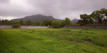 Grassy inner yard of Côn Đảo Prison on Côn Sơn Island, Vietnam, bordered by a stone wall with a lone guard tower and forested mountains under a cloudy sky