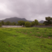 Grassy inner yard of Côn Đảo Prison on Côn Sơn Island, Vietnam, bordered by a stone wall with a lone guard tower and forested mountains under a cloudy sky