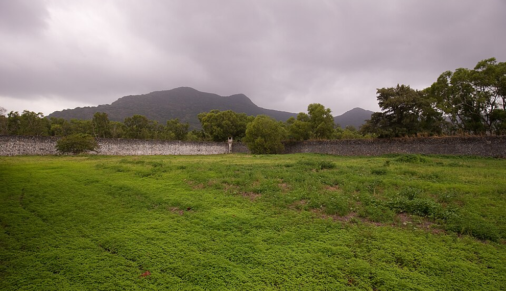 Grassy inner yard of Côn Đảo Prison on Côn Sơn Island, Vietnam, bordered by a stone wall with a lone guard tower and forested mountains under a cloudy sky