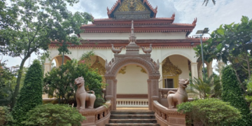 Back view of the vihara at Wat Bo pagoda in Siem Reap, showing a decorative archway with two gajasimha statues, stairway and lush green trees around the temple