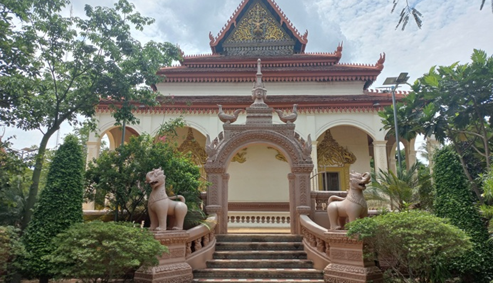 Back view of the vihara at Wat Bo pagoda in Siem Reap, showing a decorative archway with two gajasimha statues, stairway and lush green trees around the temple