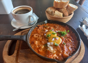 Cast-iron pan of chakchouka topped with eggs and herbs, served on a wooden board with a basket of sliced baguette and a cup of black coffee at Paris Bakery in Siem Reap.