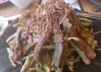 Close-up of Cambodian morning glory salad at Kraya Angkor Restaurant in Phnom Penh, featuring crispy battered morning glory topped with sliced grilled beef and golden fried shallots on a dark plate.