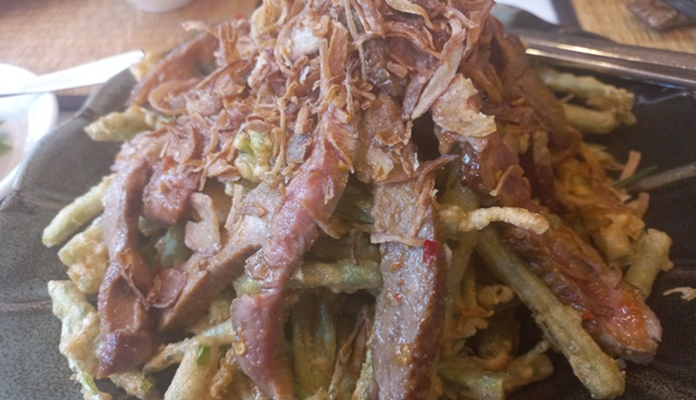 Close-up of Cambodian morning glory salad at Kraya Angkor Restaurant in Phnom Penh, featuring crispy battered morning glory topped with sliced grilled beef and golden fried shallots on a dark plate.