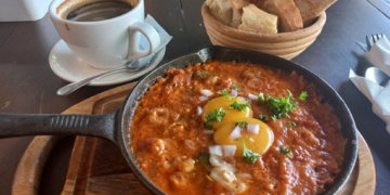 Cast-iron pan of chakchouka topped with eggs and herbs, served on a wooden board with a basket of sliced baguette and a cup of black coffee at Paris Bakery in Siem Reap.