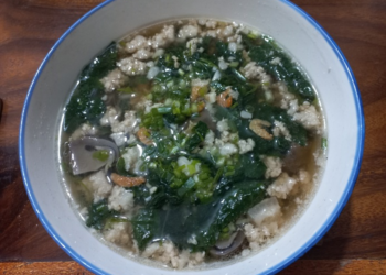 Homemade Cambodian soup in a white bowl, filled with minced pork, ivy gourd leaves, dried shrimp, and mushrooms in a clear broth on a wooden table
