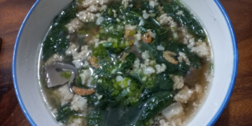 Homemade Cambodian soup in a white bowl, filled with minced pork, ivy gourd leaves, dried shrimp, and mushrooms in a clear broth on a wooden table