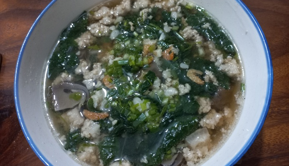 Homemade Cambodian soup in a white bowl, filled with minced pork, ivy gourd leaves, dried shrimp, and mushrooms in a clear broth on a wooden table