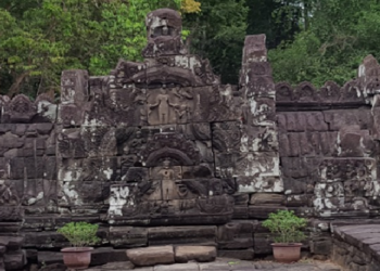 Stone naga friezes and weathered carvings at Neak Poan temple in Angkor, framed by laterite steps, potted plants, and the green edge of the surrounding baray.