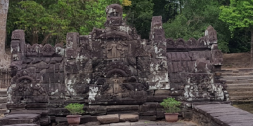Stone naga friezes and weathered carvings at Neak Poan temple in Angkor, framed by laterite steps, potted plants, and the green edge of the surrounding baray.