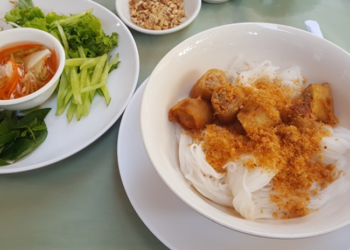 Bowl of nom banhchok Kampot rice noodles topped with dried shrimp, chopped spring rolls, and ground peanuts, served alongside a plate of fresh herbs, bean sprouts, cucumber, and dipping sauce on a pale green tablecloth.