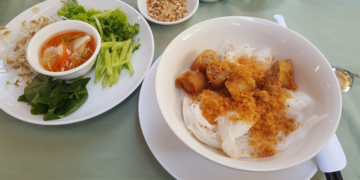 Bowl of nom banhchok Kampot rice noodles topped with dried shrimp, chopped spring rolls, and ground peanuts, served alongside a plate of fresh herbs, bean sprouts, cucumber, and dipping sauce on a pale green tablecloth.