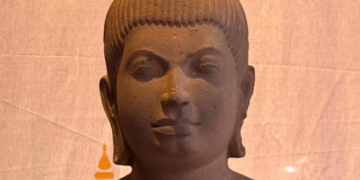 Sandstone 7th‑century Devi statue from Sambor Prei Kuk style, showing elegant Khmer feminine form with high jata hairstyle and sampot, displayed against a warm backdrop.