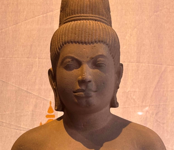 Sandstone 7th‑century Devi statue from Sambor Prei Kuk style, showing elegant Khmer feminine form with high jata hairstyle and sampot, displayed against a warm backdrop.