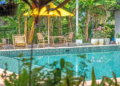 Outdoor swimming pool at Sanssouci Kep surrounded by tropical plants, bamboo loungers, and yellow umbrellas in a lush garden setting