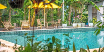 Outdoor swimming pool at Sanssouci Kep surrounded by tropical plants, bamboo loungers, and yellow umbrellas in a lush garden setting
