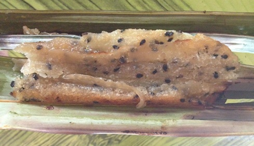 Close-up of Cambodian Nom Sleuk Chak from Kampot, a sticky rice flour cake with coconut, palm sugar, and black sesame seeds, unwrapped from nypa palm leaves on a green table.