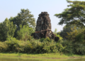 Overgrown four-faced Angkorian tower of Banteay Chhmar reflected in a tranquil baray, with Bayon-style smiling faces from the era of Jayavarman VII framed by lush Cambodian greenery.