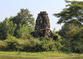 Overgrown four-faced Angkorian tower of Banteay Chhmar reflected in a tranquil baray, with Bayon-style smiling faces from the era of Jayavarman VII framed by lush Cambodian greenery.