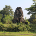 Overgrown four-faced Angkorian tower of Banteay Chhmar reflected in a tranquil baray, with Bayon-style smiling faces from the era of Jayavarman VII framed by lush Cambodian greenery.