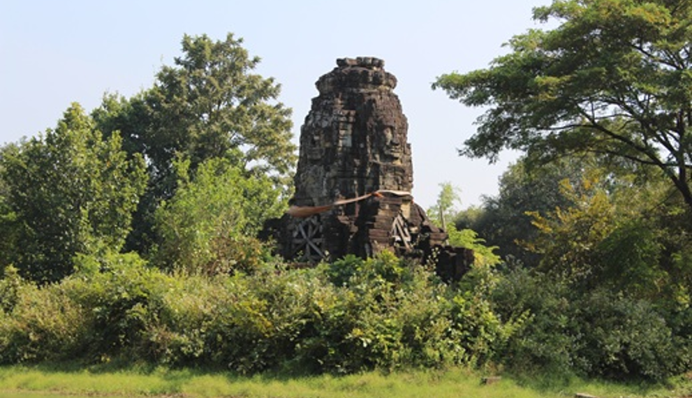 Overgrown four-faced Angkorian tower of Banteay Chhmar reflected in a tranquil baray, with Bayon-style smiling faces from the era of Jayavarman VII framed by lush Cambodian greenery.