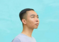 Side profile portrait of Cambodian painter Chov Theanly, wearing a light grey T‑shirt and gazing thoughtfully upward against a smooth turquoise background
