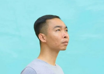 Side profile portrait of Cambodian painter Chov Theanly, wearing a light grey T‑shirt and gazing thoughtfully upward against a smooth turquoise background