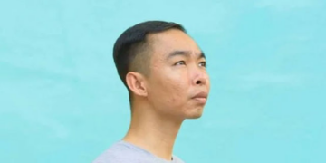 Side profile portrait of Cambodian painter Chov Theanly, wearing a light grey T‑shirt and gazing thoughtfully upward against a smooth turquoise background