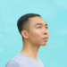 Side profile portrait of Cambodian painter Chov Theanly, wearing a light grey T‑shirt and gazing thoughtfully upward against a smooth turquoise background