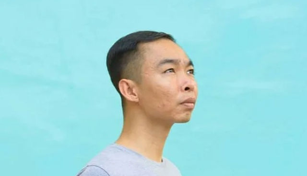 Side profile portrait of Cambodian painter Chov Theanly, wearing a light grey T‑shirt and gazing thoughtfully upward against a smooth turquoise background