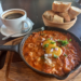 Cast-iron pan of chakchouka topped with eggs and herbs, served on a wooden board with a basket of sliced baguette and a cup of black coffee at Paris Bakery in Siem Reap.