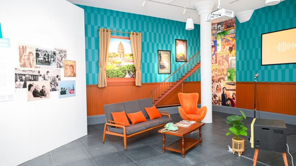Reconstitution d’un salon cambodgien des années du Sangkum avec canapé et fauteuil orange, télévision vintage et décor mural coloré au Musée SOSORO