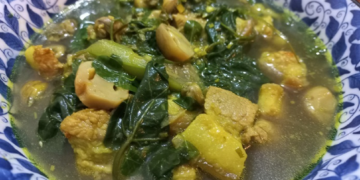 Close up of a bowl of samla prahae, traditional Cambodian vegetable meat soup with pumpkin, leafy greens and fermented fish in a clear lemongrass broth.