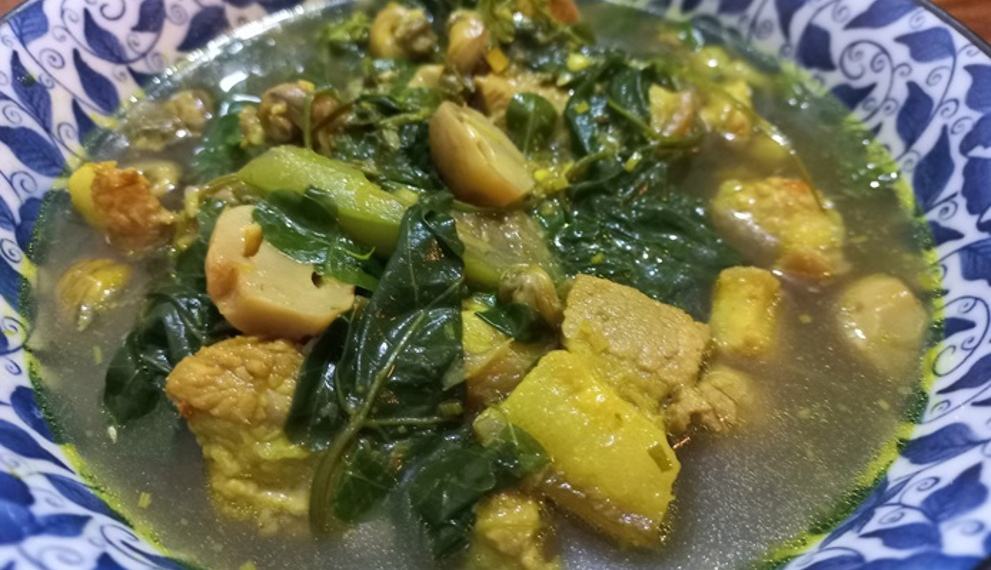 Close up of a bowl of samla prahae, traditional Cambodian vegetable meat soup with pumpkin, leafy greens and fermented fish in a clear lemongrass broth.