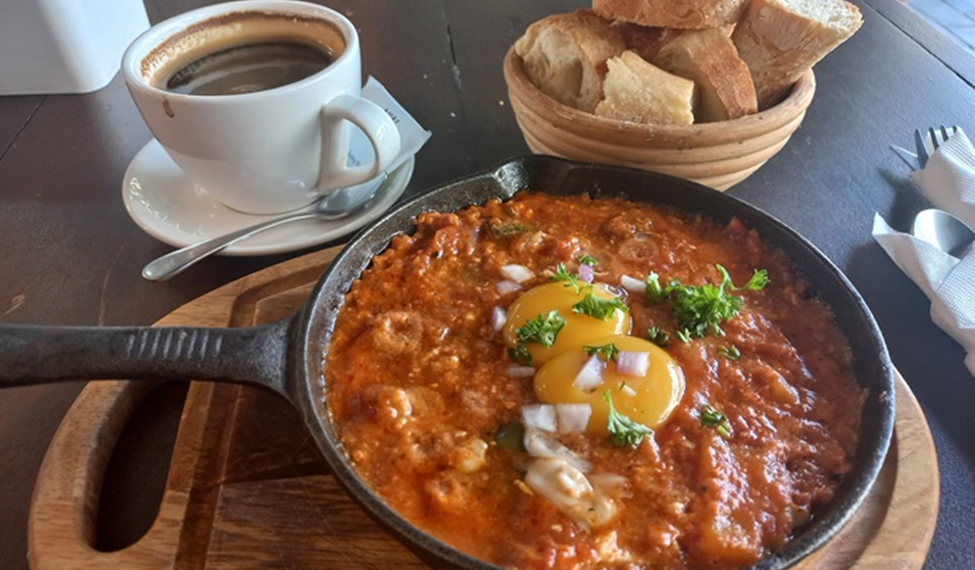 Cast-iron pan of chakchouka topped with eggs and herbs, served on a wooden board with a basket of sliced baguette and a cup of black coffee at Paris Bakery in Siem Reap.