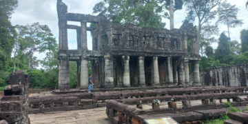 Two storied stone pavilion with round columns at Preah Khan temple in Angkor, surrounded by lush trees and ancient terraces, Siem Reap, Cambodia.