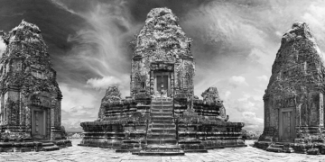 Black‑and‑white panoramic view of Pre Rup’s upper terrace at Angkor, showing the central stepped sandstone prasat flanked by two smaller brick towers under a dramatic cloudy sky.
