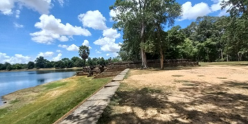 Stone terrace and shaded trees on the western edge of Sras Srang, with the large Angkor reservoir stretching out under blue sky and scattered clouds at midday.