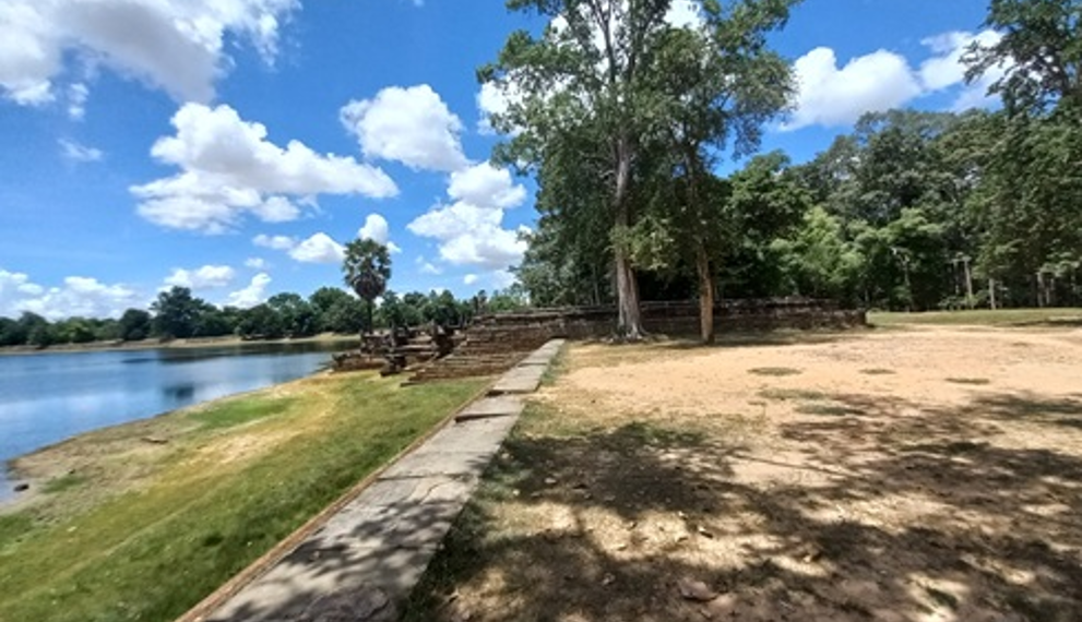 Stone terrace and shaded trees on the western edge of Sras Srang, with the large Angkor reservoir stretching out under blue sky and scattered clouds at midday.