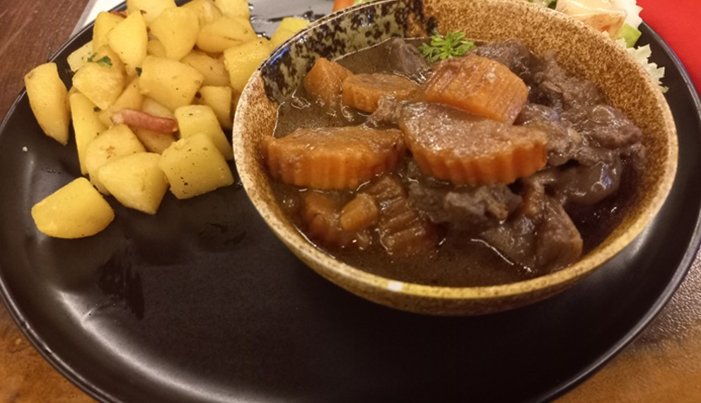 Close-up of boeuf-carottes with braised beef cheek and carrots, served in a rustic bowl alongside sautéed diced potatoes and fresh salad on a black plate at Graines de Piaf French bistrot in Siem Reap.