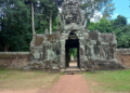 Stone entrance gate on the east side of Banteay Kdei temple, featuring a tall tower with a serene sculpted face, weathered sandstone and lichens, framed by dense green trees and blue sky, with a reddish dirt path leading through the gateway into the forested interior.