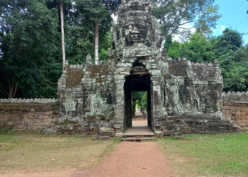 Stone entrance gate on the east side of Banteay Kdei temple, featuring a tall tower with a serene sculpted face, weathered sandstone and lichens, framed by dense green trees and blue sky, with a reddish dirt path leading through the gateway into the forested interior.