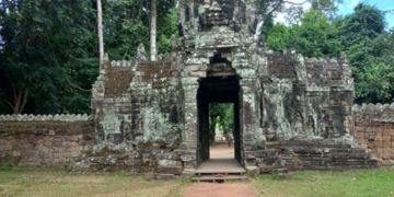 Stone entrance gate on the east side of Banteay Kdei temple, featuring a tall tower with a serene sculpted face, weathered sandstone and lichens, framed by dense green trees and blue sky, with a reddish dirt path leading through the gateway into the forested interior.