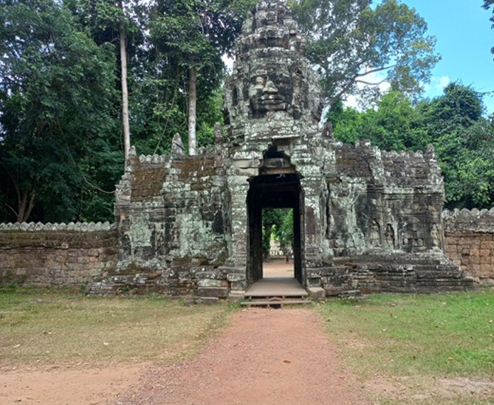Stone entrance gate on the east side of Banteay Kdei temple, featuring a tall tower with a serene sculpted face, weathered sandstone and lichens, framed by dense green trees and blue sky, with a reddish dirt path leading through the gateway into the forested interior.
