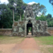 Stone entrance gate on the east side of Banteay Kdei temple, featuring a tall tower with a serene sculpted face, weathered sandstone and lichens, framed by dense green trees and blue sky, with a reddish dirt path leading through the gateway into the forested interior.