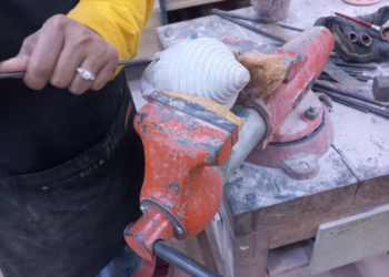 Close-up of a craftsman in a yellow shirt using a hand tool to carve a small stone Buddha head clamped in an orange vise on a dusty workbench
