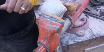 Close-up of a craftsman in a yellow shirt using a hand tool to carve a small stone Buddha head clamped in an orange vise on a dusty workbench