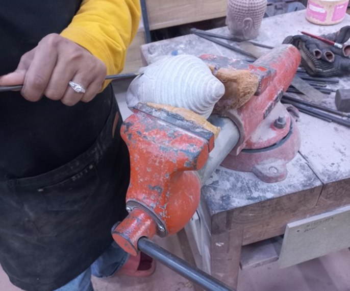 Close-up of a craftsman in a yellow shirt using a hand tool to carve a small stone Buddha head clamped in an orange vise on a dusty workbench